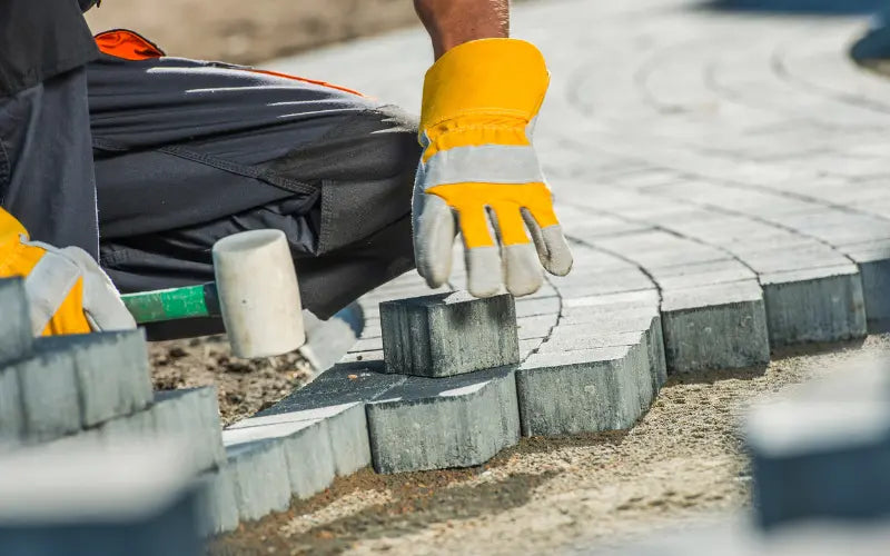 Landscaper knelt down with hammer in hand laying block paving slabs, wearing safety gloves
